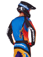 Motocross rider viewed from the back wearing the Alpinestars 2025 Fluid Wurx Gear Set, multicolor jersey with blue back and black sleeves featuring white logos, matching orange and blue pants, black and blue helmet with orange goggles, coordinated off-road racing outfit.