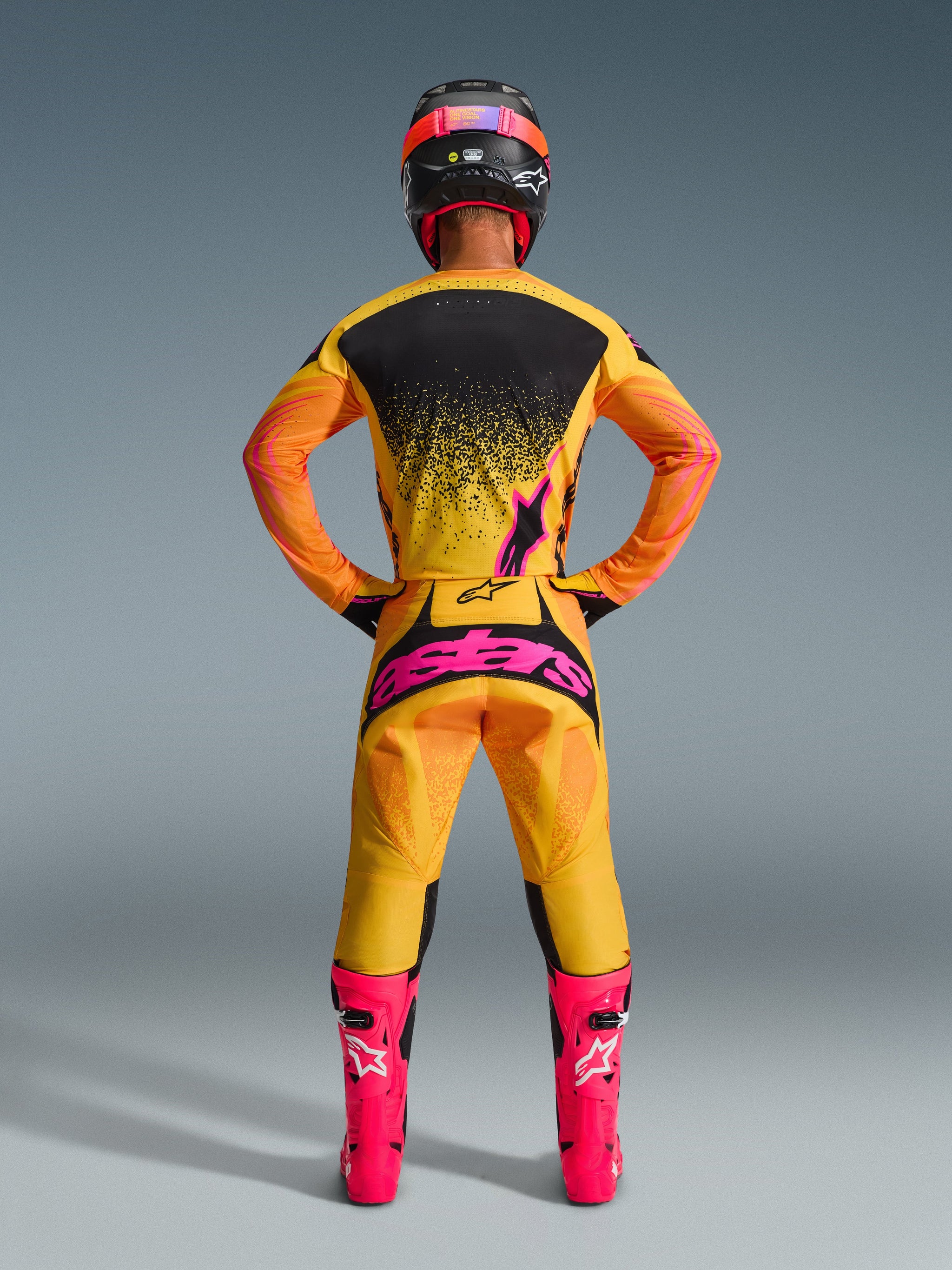 Motocross rider from the back wearing the Alpinestars 2026 Techstar Nomur Jersey in Orange Yellow Pink, coordinated racing gear including matching pants with bold pink branding and bright pink boots, athletic race-fit for advanced level off-road performance.