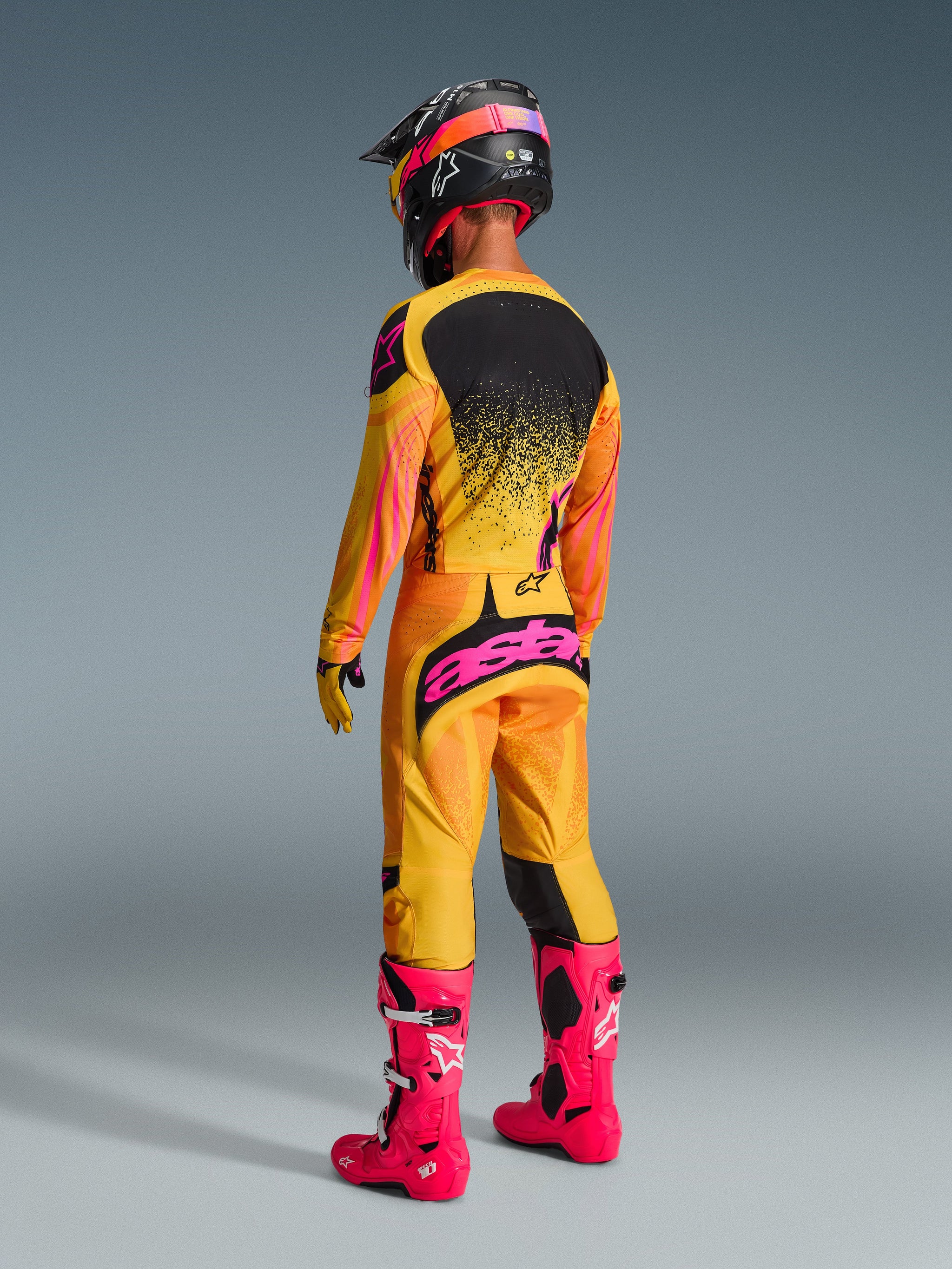 Motocross rider from the back wearing Alpinestars 2026 Techstar Nomur Jersey, Orange Yellow and Pink, matching racing pants with pink branding, bright pink boots, and a coordinated black and pink helmet, full off-road competition gear set