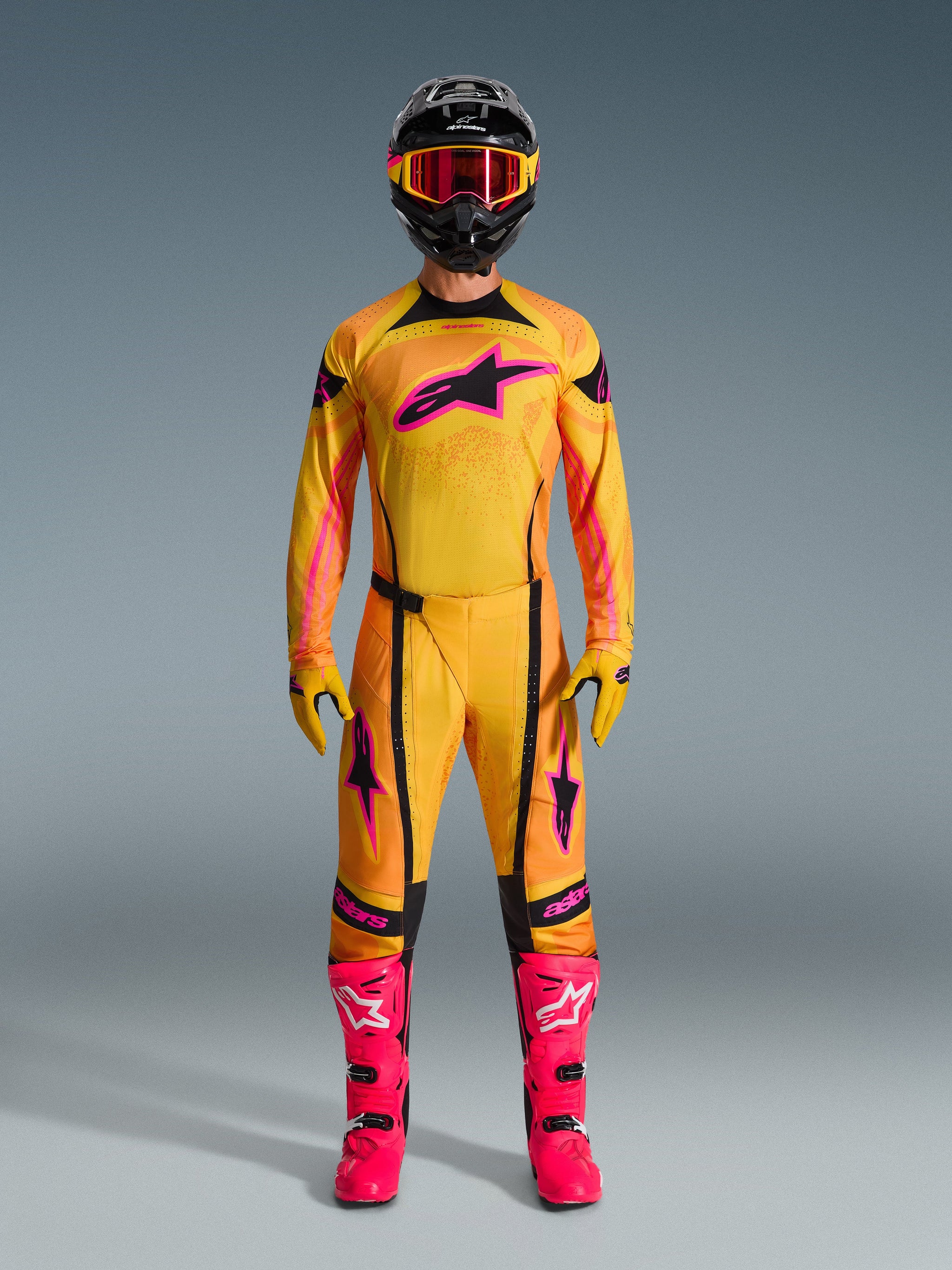 Motocross rider wearing the 2026 Techstar Nomur Jersey in Orange, Yellow, and Pink, coordinated with matching motocross pants, a black helmet with pink goggles, and vibrant pink boots, athletic race-fit gear for off-road competition