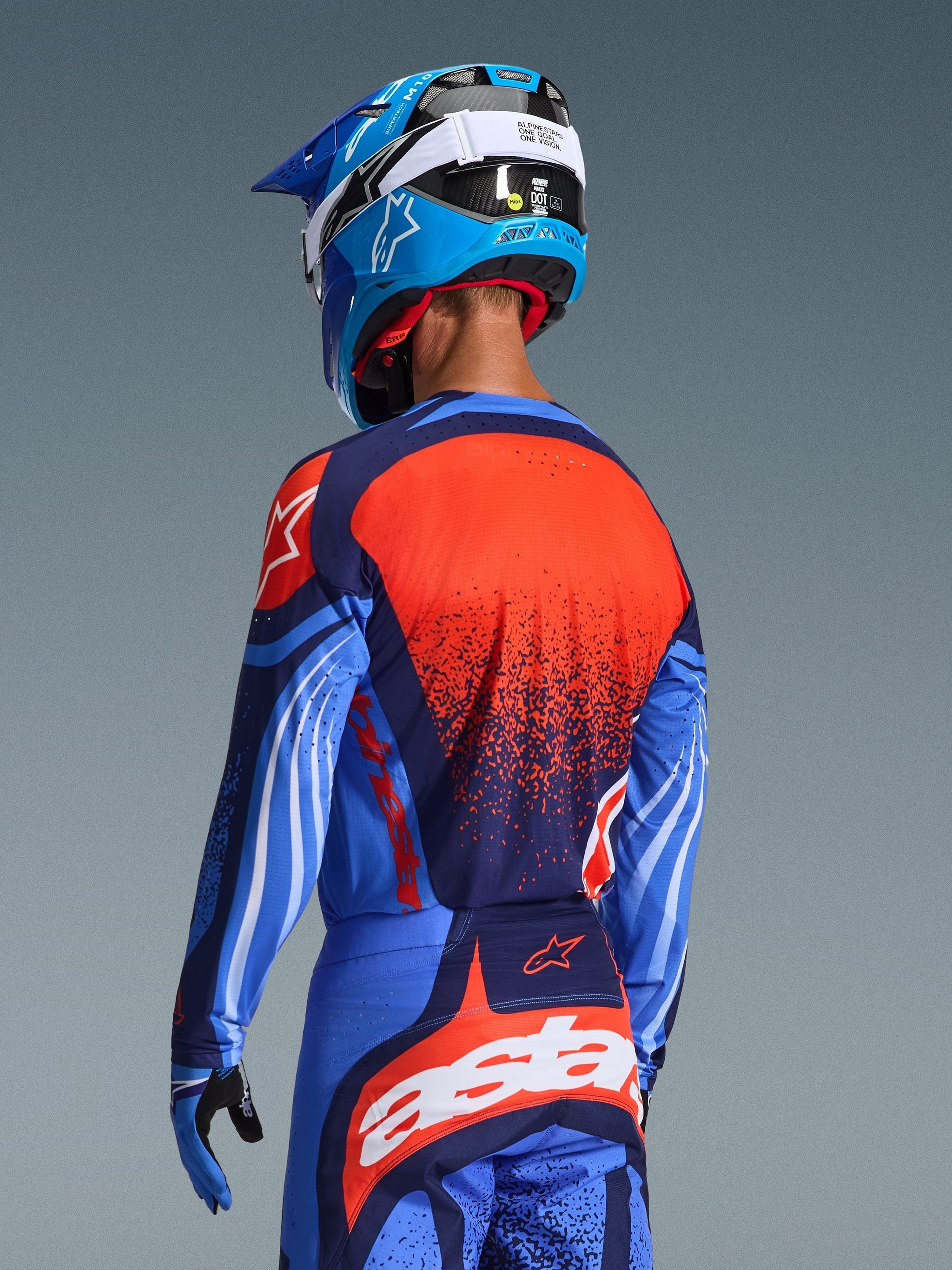 Motocross rider from behind wearing the 2026 Techstar Nomur Jersey, Dark Navy with Orange and UCLA Blue accents, featuring a large orange textured graphic on the back, paired with matching motocross pants and a blue racing helmet, coordinated professional gear for off-road riding.