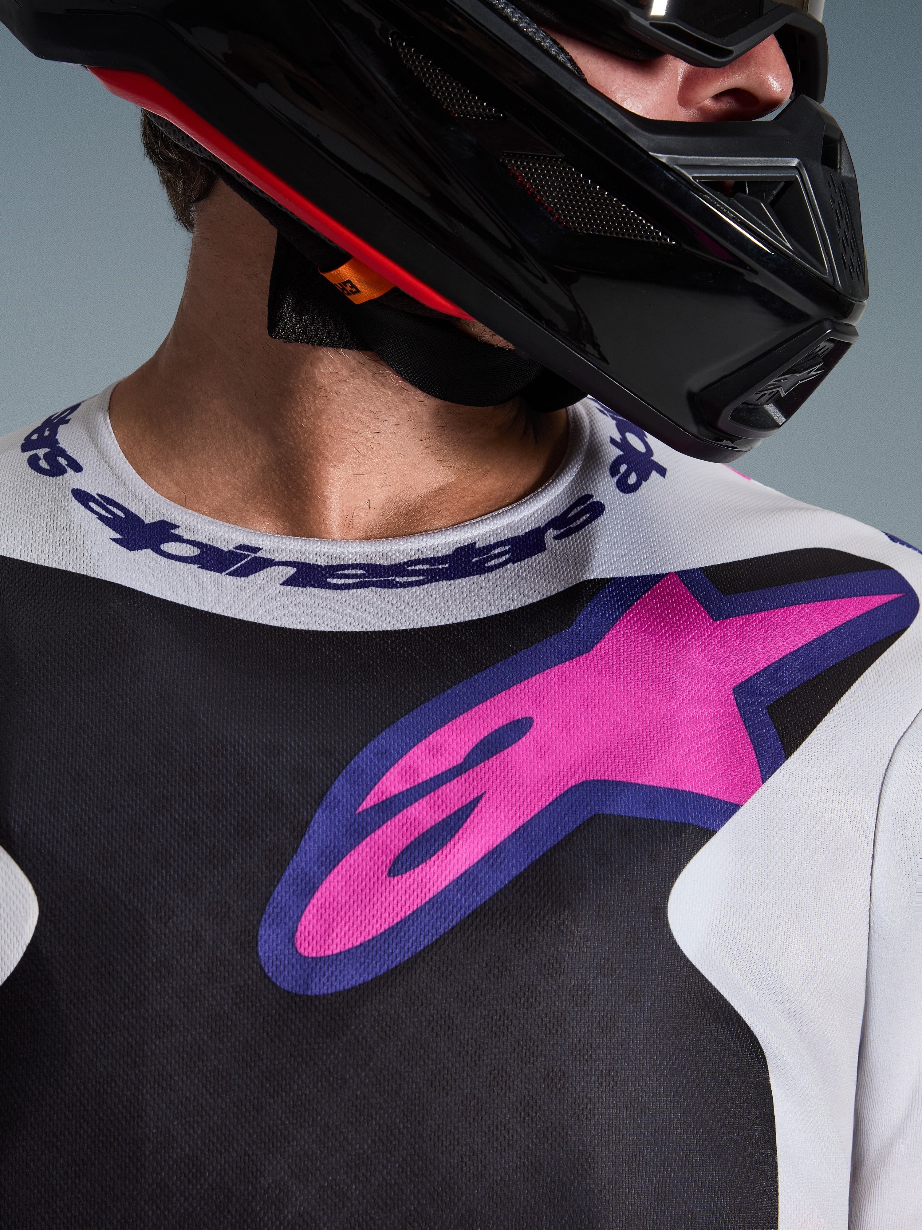 Alpinestars 2026 Fluid Grid Jersey, Motocross Jersey, Black Light Gray and Purple, close-up of chest and neck area featuring large pink star logo, branded sport collar, worn with black helmet, performance off-road gear