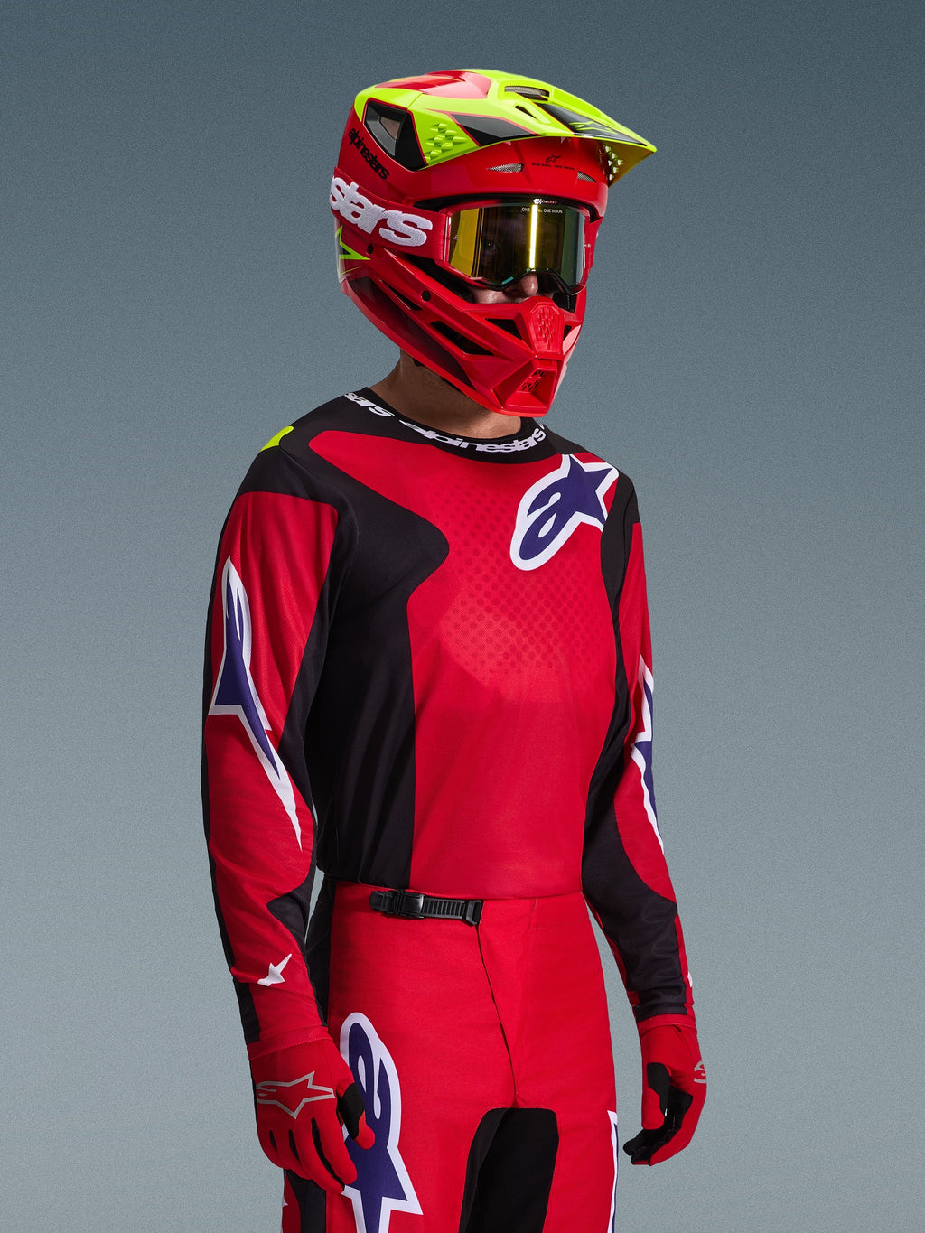 Motocross rider wearing Alpinestars 2026 Fluid Grid Gear Set, Red and Black jersey with white and blue logo accents, matching Red and Black pants, Red helmet with yellow highlights and gold goggles, coordinated off-road racing gear