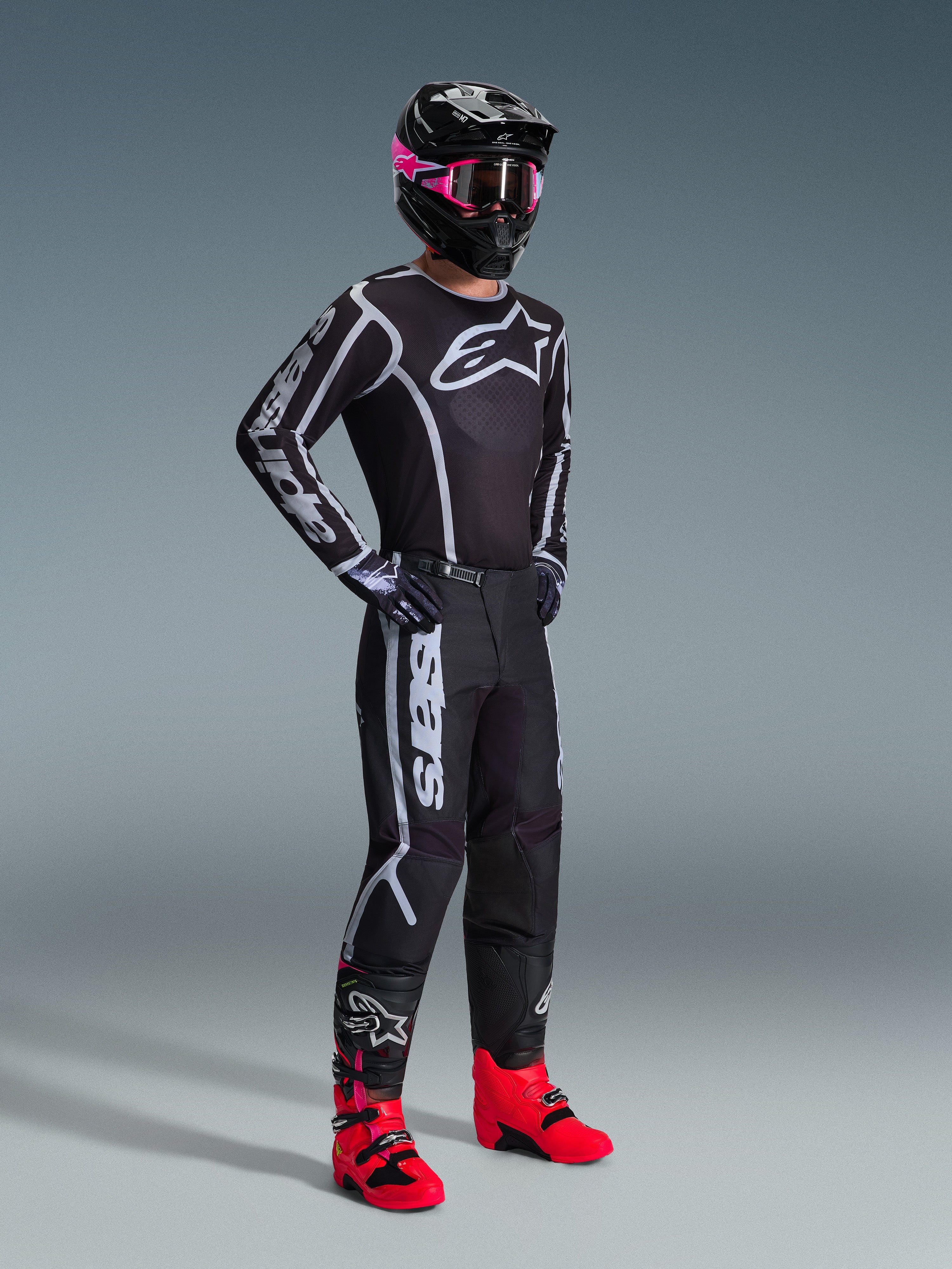 Alpinestars 2026 Fluid Apex Gear Set, motocross rider wearing a black jersey and pants with white and grey graphic accents, black helmet with pink goggles, and bright pink racing boots, coordinated professional motocross outfit