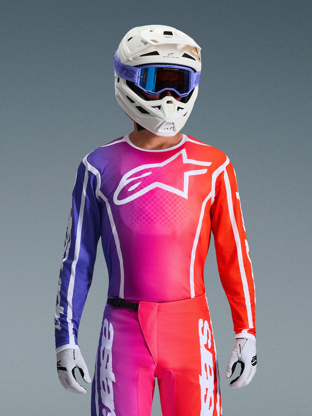 Motocross rider wearing the 2026 Fluid Apex Jersey, vibrant purple, pink, and orange gradient design with a large white Alpinestars logo on the chest, coordinated with a white helmet, purple goggles, and white gloves for a complete off-road racing look.