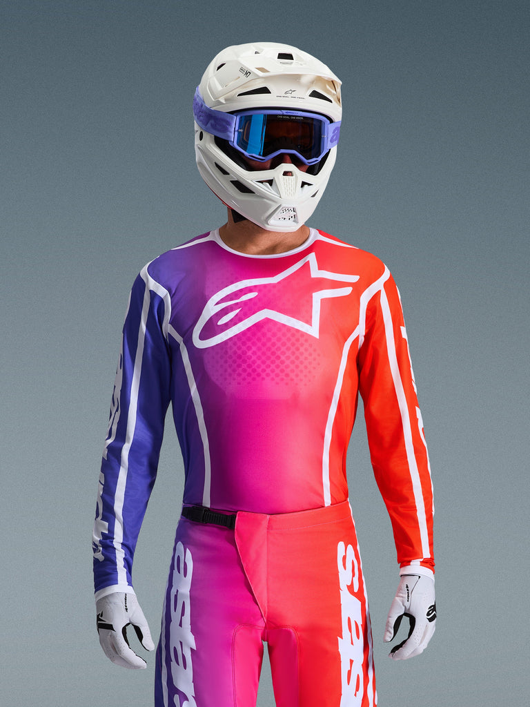 Motocross rider wearing the 2026 Fluid Apex Jersey, vibrant purple, pink, and orange gradient design with a large white Alpinestars logo on the chest, coordinated with a white helmet, purple goggles, and white gloves for a complete off-road racing look.