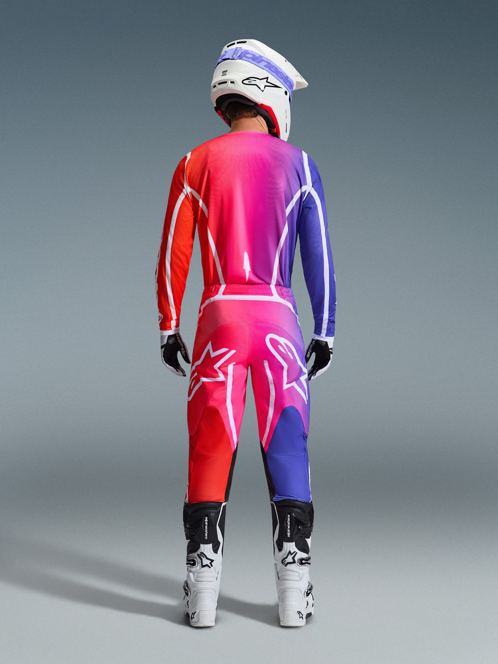 Alpinestars 2026 Fluid Apex Jersey and matching pants, Motocross gear set, Multicolor red and purple gradient, rear view of a rider wearing white and black boots and a white helmet, featuring bold white logos and geometric patterns for off-road racing.