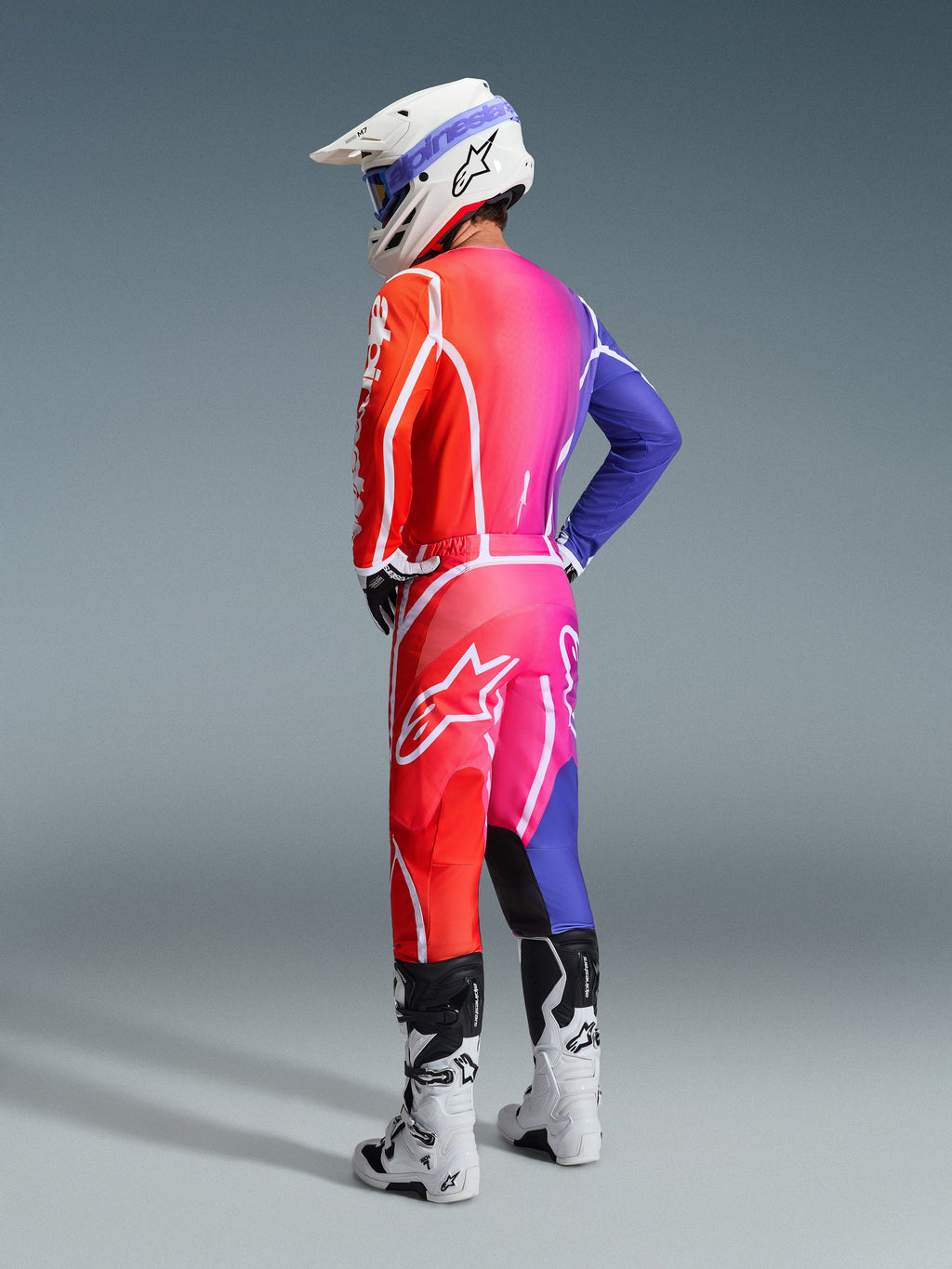 Motocross rider wearing the Alpinestars 2026 Fluid Apex Gear Set, vibrant gradient transition from orange to pink and purple with bold white branding and star logos, paired with a white helmet, blue goggles, and white boots, seen from a rear three-quarter angle in a studio setting.