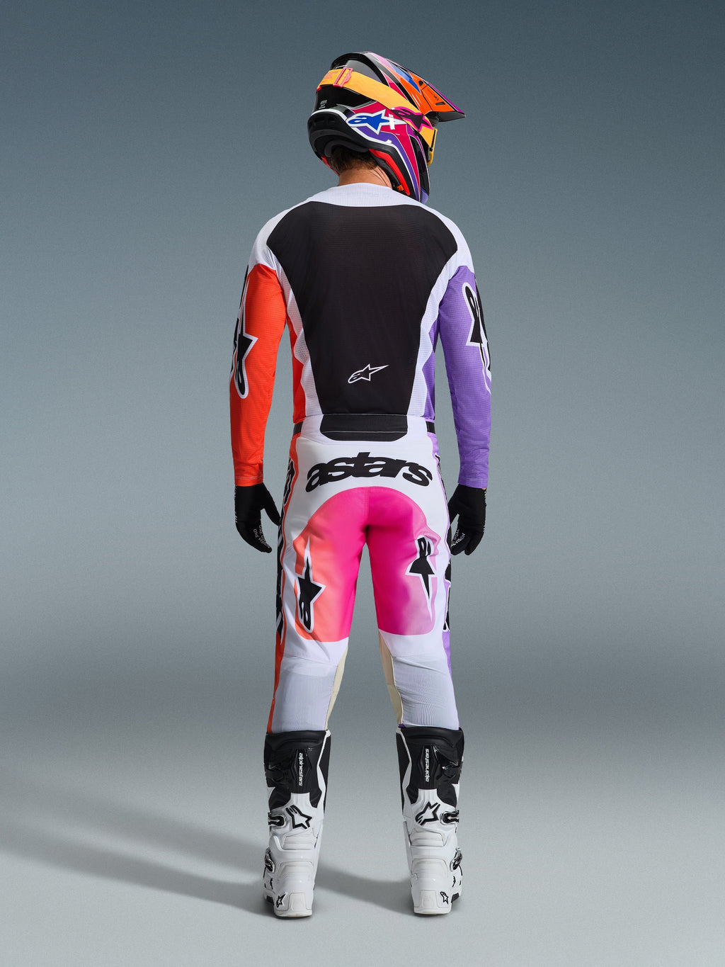 Rear view of a motocross rider wearing the Alpinestars 2026 Racer Air Portl Pants in Multicolor, featuring white fabric with pink and orange gradient panels and black branding, styled with a matching black jersey with purple and orange sleeves, white racing boots, and a vibrant multicolored helmet.