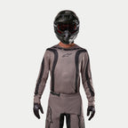 Motocross rider wearing the Alpinestars 2024 Fluid Lurv Jersey in Mud Black, an earthy grey-brown fabric with black graphic stripes and a star logo on the chest, paired with a black helmet, red goggles, and black gloves for a coordinated off-road racing look