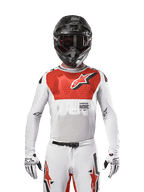 Motocross rider wearing Alpinestars 2024 Supertech Ward Jersey in White Hot Orange, white stretch mesh jersey with a prominent red chest graphic and logo, black helmet with dark goggles, white gloves, professional-level gear for off-road performance