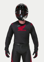 Motocross rider wearing the Alpinestars Honda Racer Iconic Jersey in Black and Red, featuring a large red Honda wing logo on the chest and white Astar logo, paired with a matching black helmet, goggles, and gloves for a complete off-road racing kit