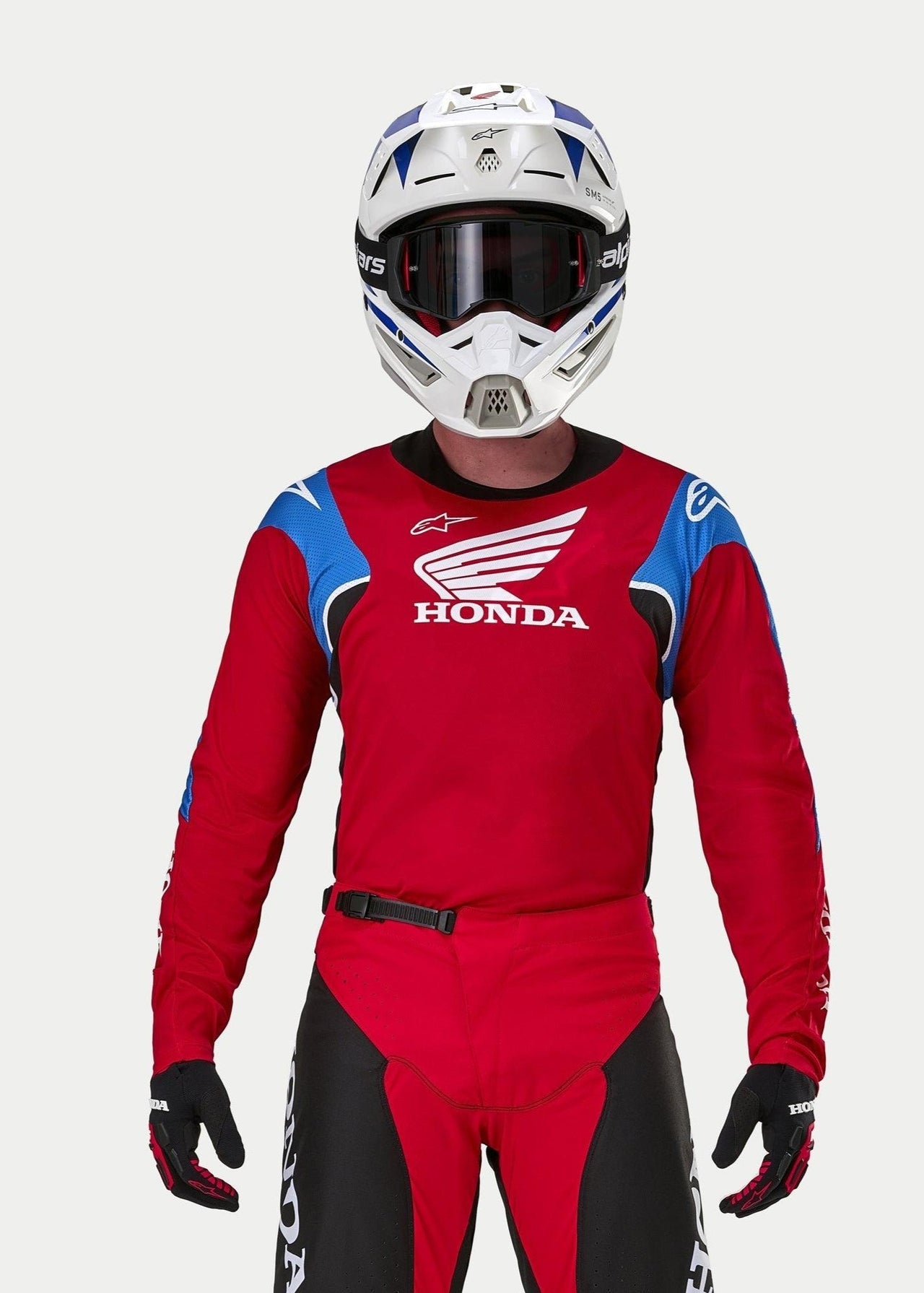 Motocross rider wearing the Alpinestars Honda Racer Iconic Jersey, Bright Red with blue and white accents and Honda branding, paired with a white and blue helmet, dark goggles, and matching red and black motocross pants, performance gear for off-road riding