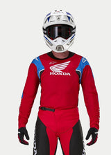 Motocross rider wearing the Alpinestars Honda Racer Iconic Jersey, Bright Red with blue and white accents and Honda branding, paired with a white and blue helmet, dark goggles, and matching red and black motocross pants, performance gear for off-road riding