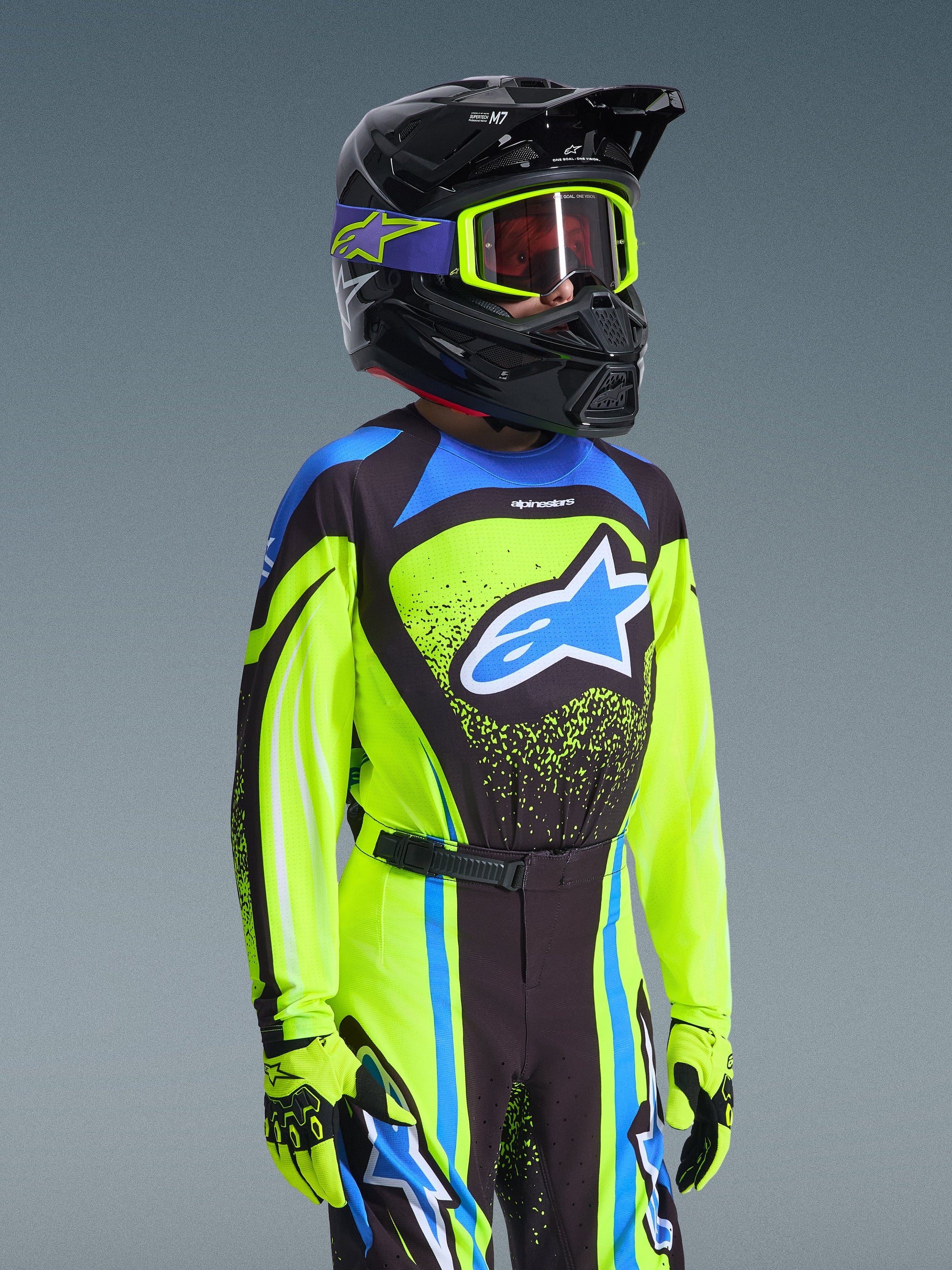 Youth motocross rider wearing Alpinestars 2026 Youth Supertech Nomur Jersey in Yellow Fluo and Dark Navy, featuring a blue Astars logo, matching racing pants and gloves, and a black helmet with fluo yellow goggles, professional off-road motocross gear.