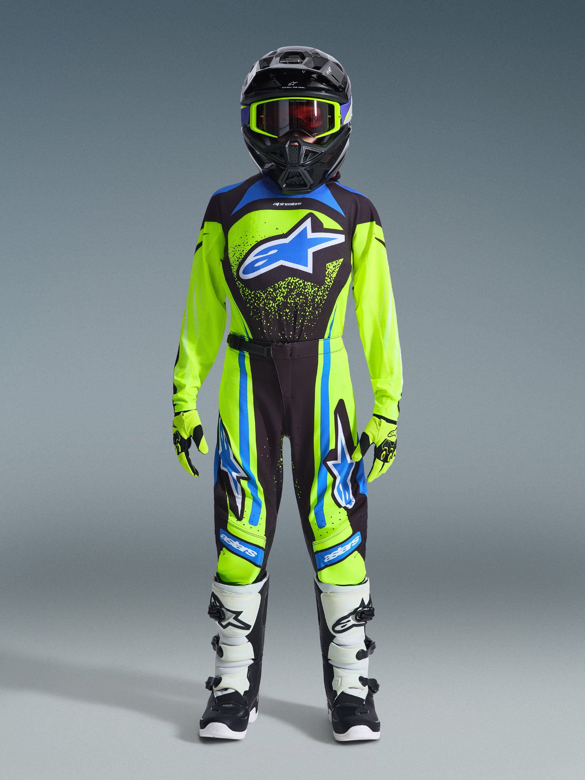 Youth motocross rider wearing complete racing gear, 2026 Youth Supertech Nomur Jersey in neon yellow and dark navy with a blue Alpinestars logo, matching pants, white and black boots, neon yellow gloves, and a black helmet with goggles, professional off-road racing outfit.