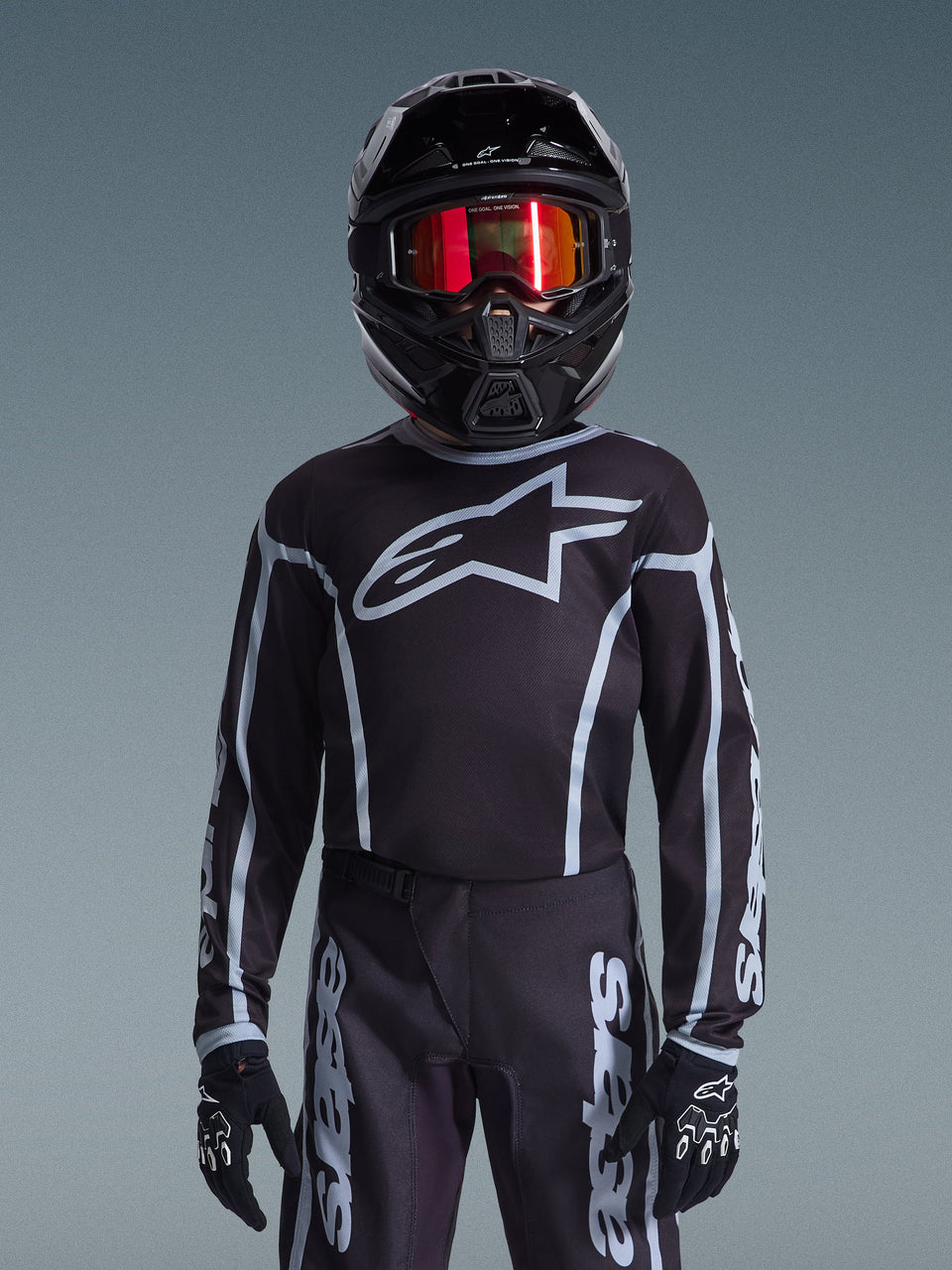 Youth motocross rider wearing Alpinestars 2026 Youth Fluid Apex Jersey in Black and Gray, matching pants, black helmet with red reflective goggles, and black gloves, coordinated off-road racing gear.
