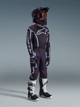 Youth motocross rider wearing the 2026 Youth Fluid Apex Jersey in Black Gray, matching black and gray pants with white graphics, black helmet with red-tinted goggles, black gloves, and white and black motocross boots, complete off-road racing kit.
