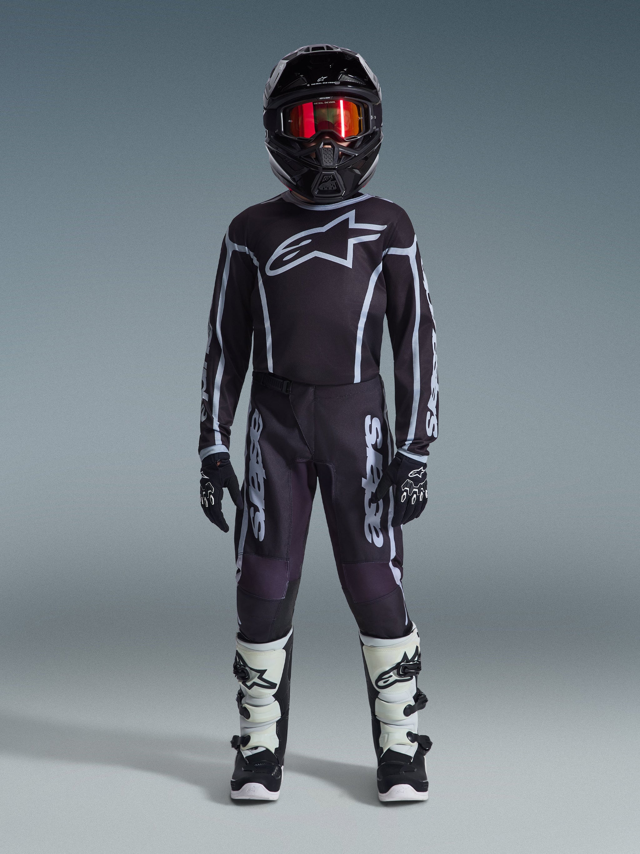 Young motocross rider wearing the Alpinestars 2026 Youth Fluid Apex Jersey in Black and Gray, coordinated with matching pants, black helmet with red iridescent goggles, black and white gloves, and motocross boots, complete gear set for youth off-road racing