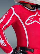 Close-up of Alpinestars 2026 Youth Fluid Apex Jersey in Red and White, featuring a large white chest logo and geometric sleeve accents, worn by a rider with black motocross gloves and a white helmet.