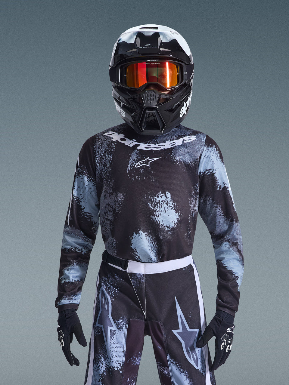 Alpinestars 2026 Youth Fluid Lahnd Jersey, Motocross Jersey, Iron Camo, worn by a youth rider with matching pants, black and white helmet with orange goggles, and black gloves, featuring a splattered grey and black design with white branding, complete off-road racing gear for young riders.