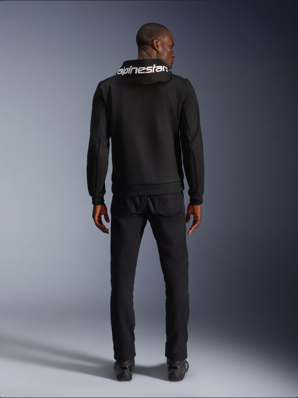 Alpinestars Chrome V2 Sport Hoodie, Motorcycle Hoodie, Black, back view of model wearing black tech fleece hoodie with white Alpinestars logo on the hood, paired with black pants and boots, casual urban riding style