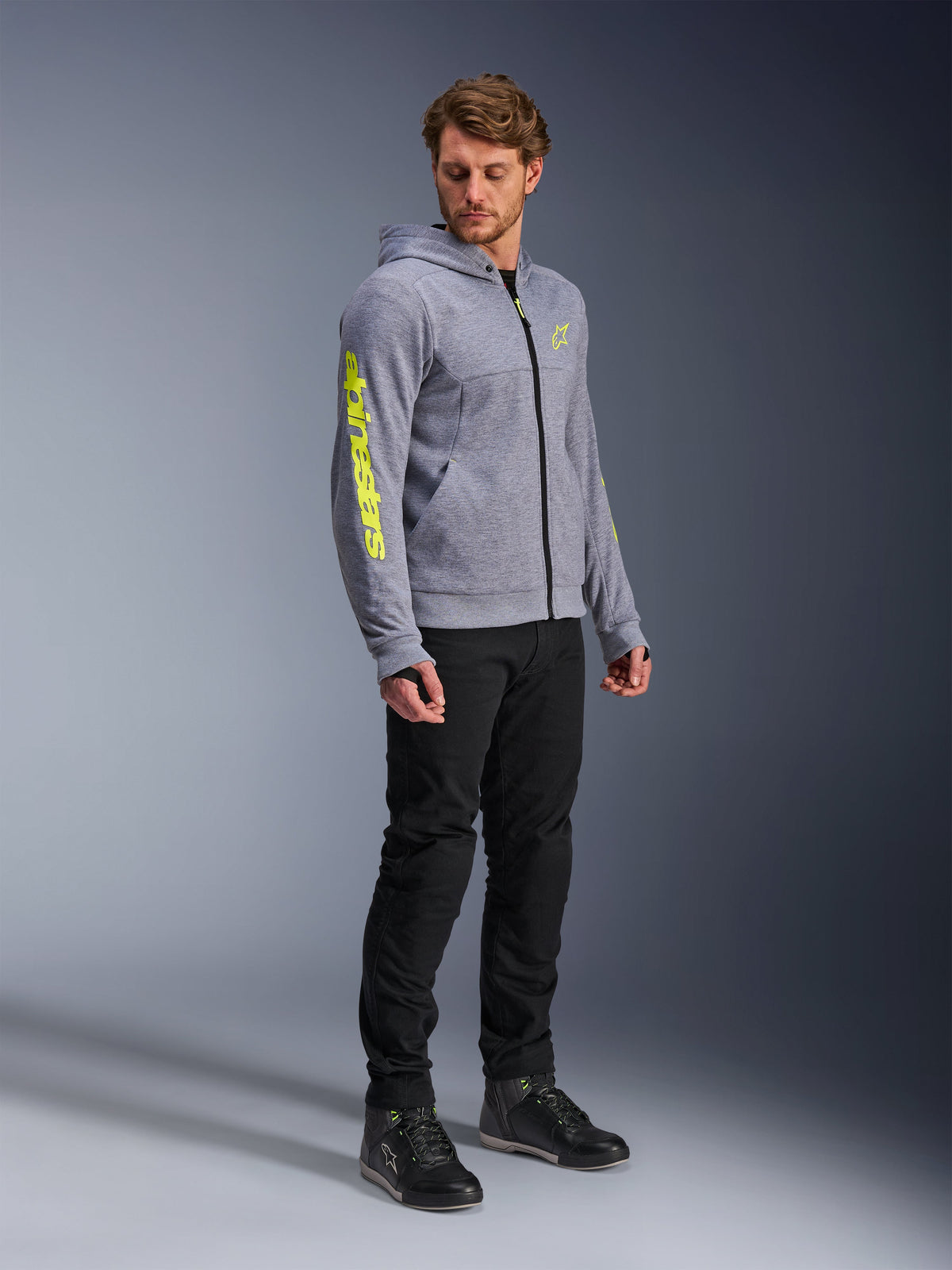 Alpinestars Chrome V2 Sport Hoodie, Gray Heather and Fluo Yellow, worn by a male model with black pants and black riding shoes, featuring bold yellow branding on the sleeve and chest, casual urban motorcycle riding gear