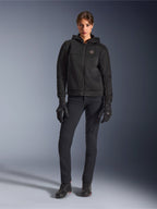 Female model wearing the Alpinestars Stella Chrome Superairflow Hoodie in Black, paired with black technical riding pants and gloves, featuring a 3D mesh textile construction and copper-toned chest logo, urban sport motorcycle apparel for women.