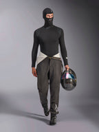 Alpinestars ZX Evo V3 Balaclava, Black, worn by a model in a full racing base layer outfit including a matching long-sleeve top and grey racing pants, holding a carbon fiber helmet with an iridescent visor, professional auto racing gear