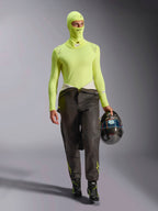 Male racing driver wearing Alpinestars ZX Evo V3 Balaclava in Yellow Fluo, matching long-sleeve base layer top, dark racing pants, and black racing boots, holding a carbon fiber helmet, professional automotive racing gear