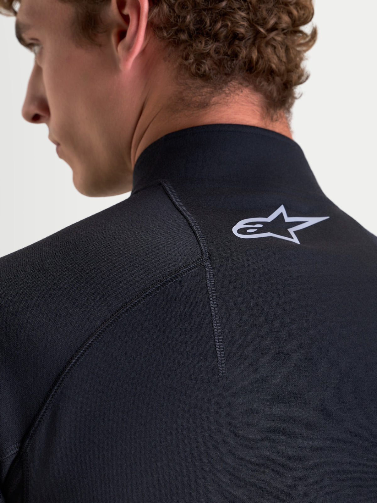 Alpinestars AT-8 Merino Top Layer, men's adventure motorcycle base layer in black, close-up back view on a model showing raglan sleeve construction, streamlined collar, and white Alpinestars logo detail