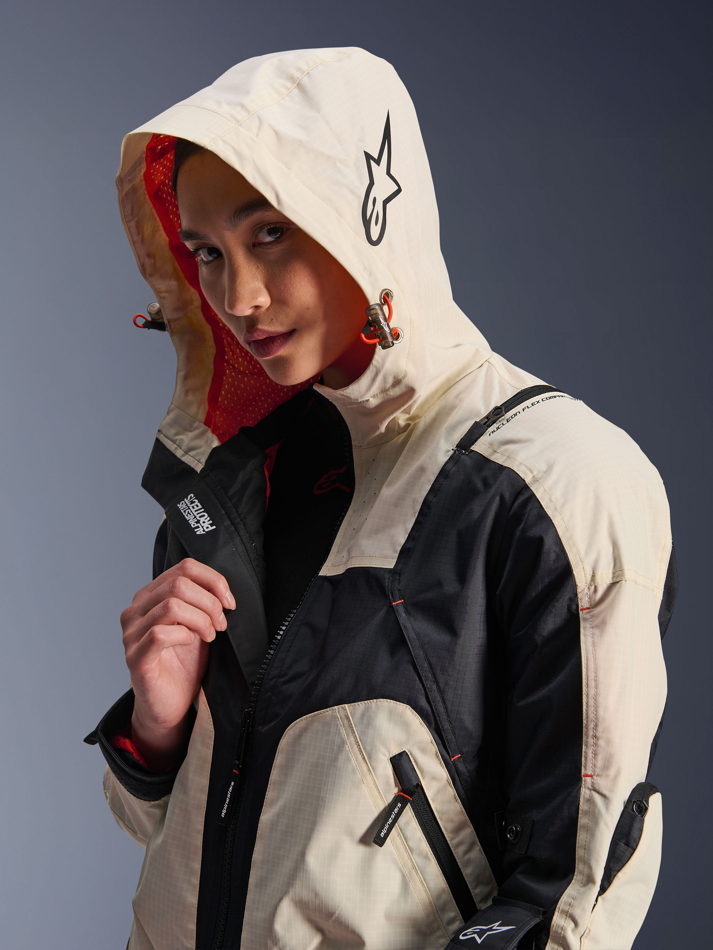 Alpinestars Molly Air Jacket in Black Almond, worn by a female model with the hood up, featuring cream and black color-blocking, a vibrant orange mesh lining, and technical urban design elements including a star logo and zip details.