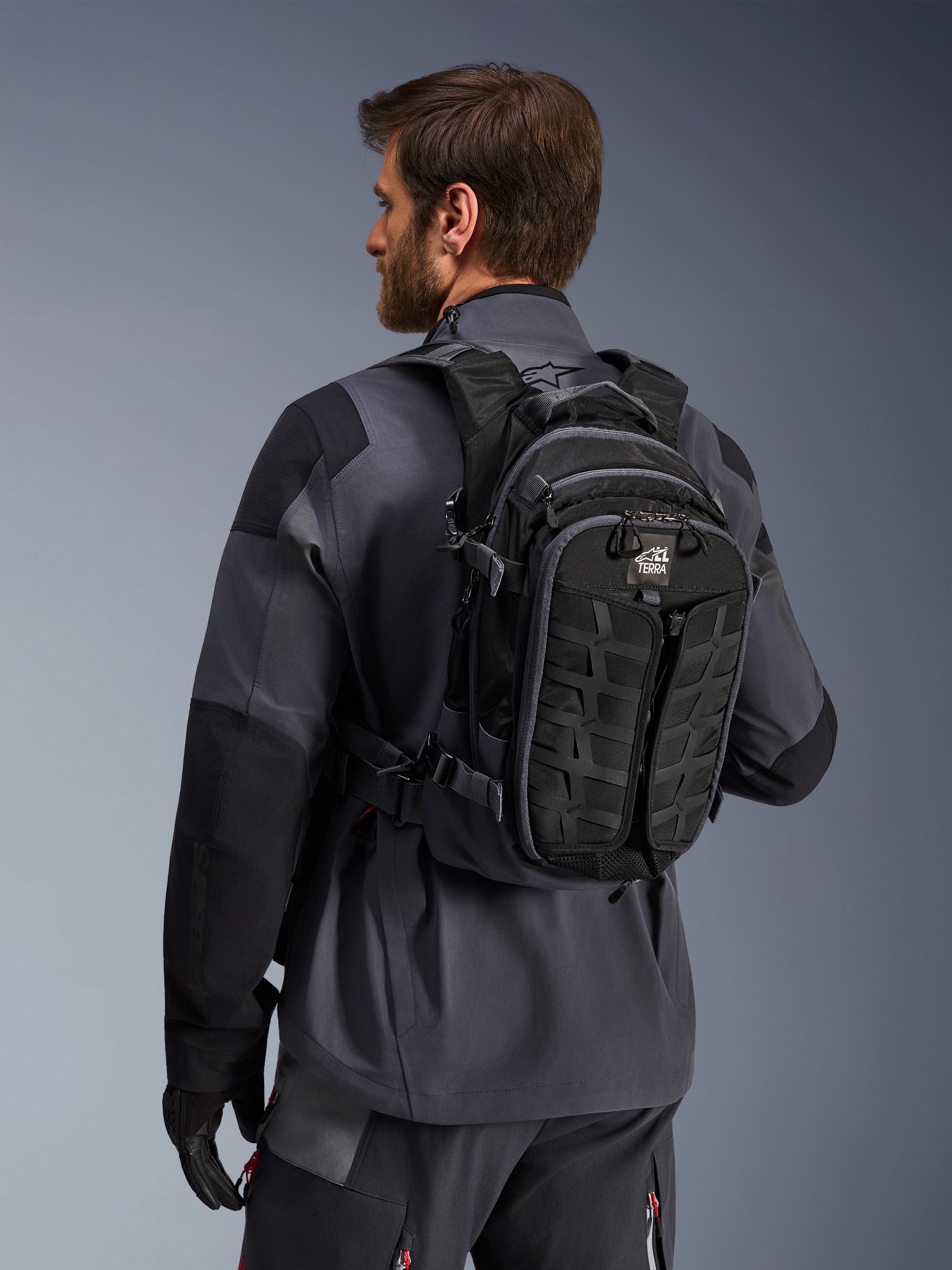 Alpinestars AT-15 Back Pack, Adventure Motorcycle Backpack, Black and Deep Gray, worn by a rider in a gray jacket, featuring a technical design with MOLLE webbing and 'TERRA' branding, ergonomic off-road gear
