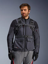 Alpinestars AT-15 Back Pack harness system, Black Deep Gray, front view worn by a rider over a gray motorcycle jacket, featuring magnetic chest lock and secure waist strap, paired with black gloves for adventure motorcycling