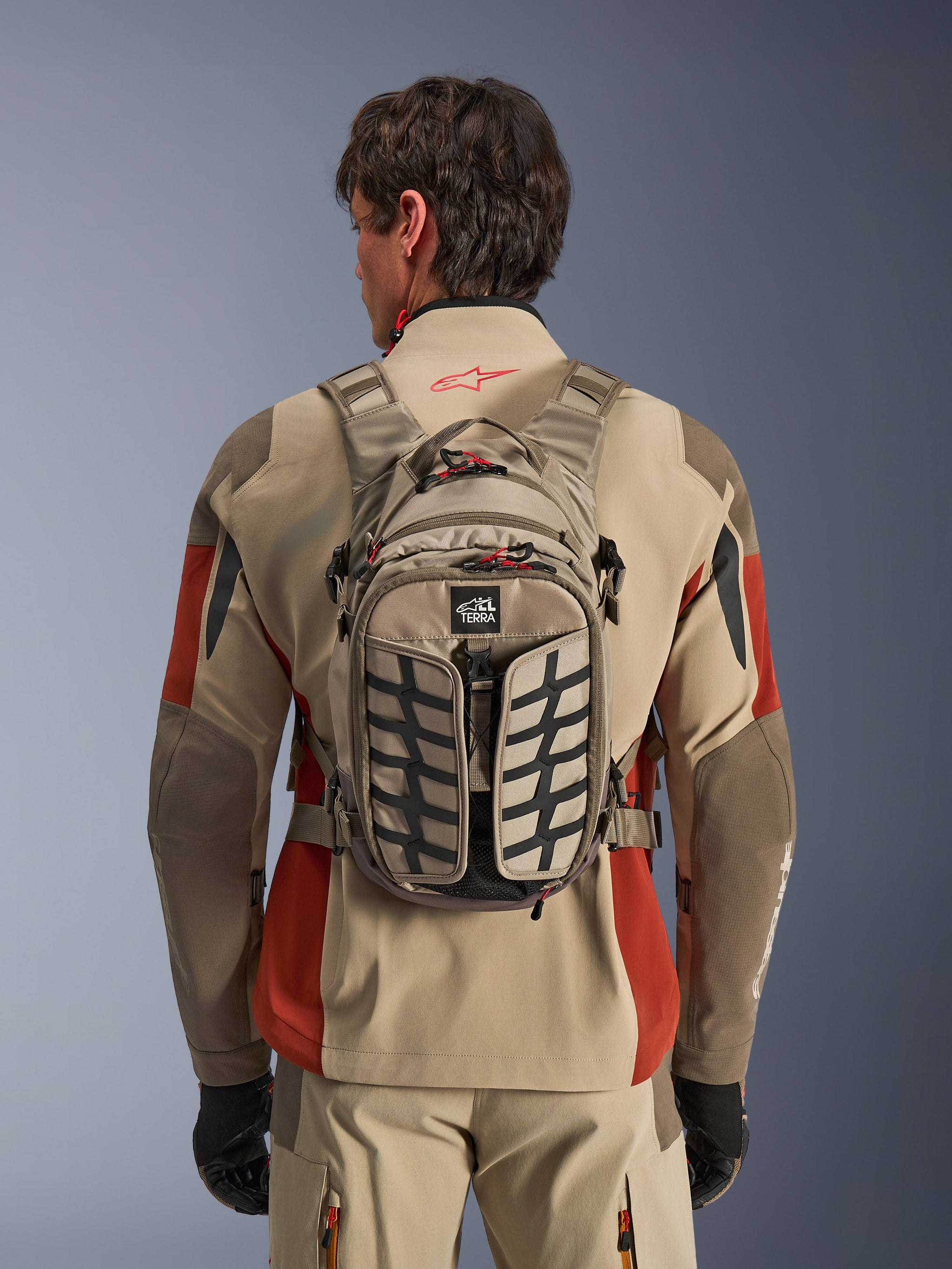 Alpinestars AT-15 Back Pack, motorcycle adventure backpack, beige and black, tire-tread pattern design, red zipper pulls, TERRA label, worn by rider with beige and red jacket, compact size with adjustable straps and buckles