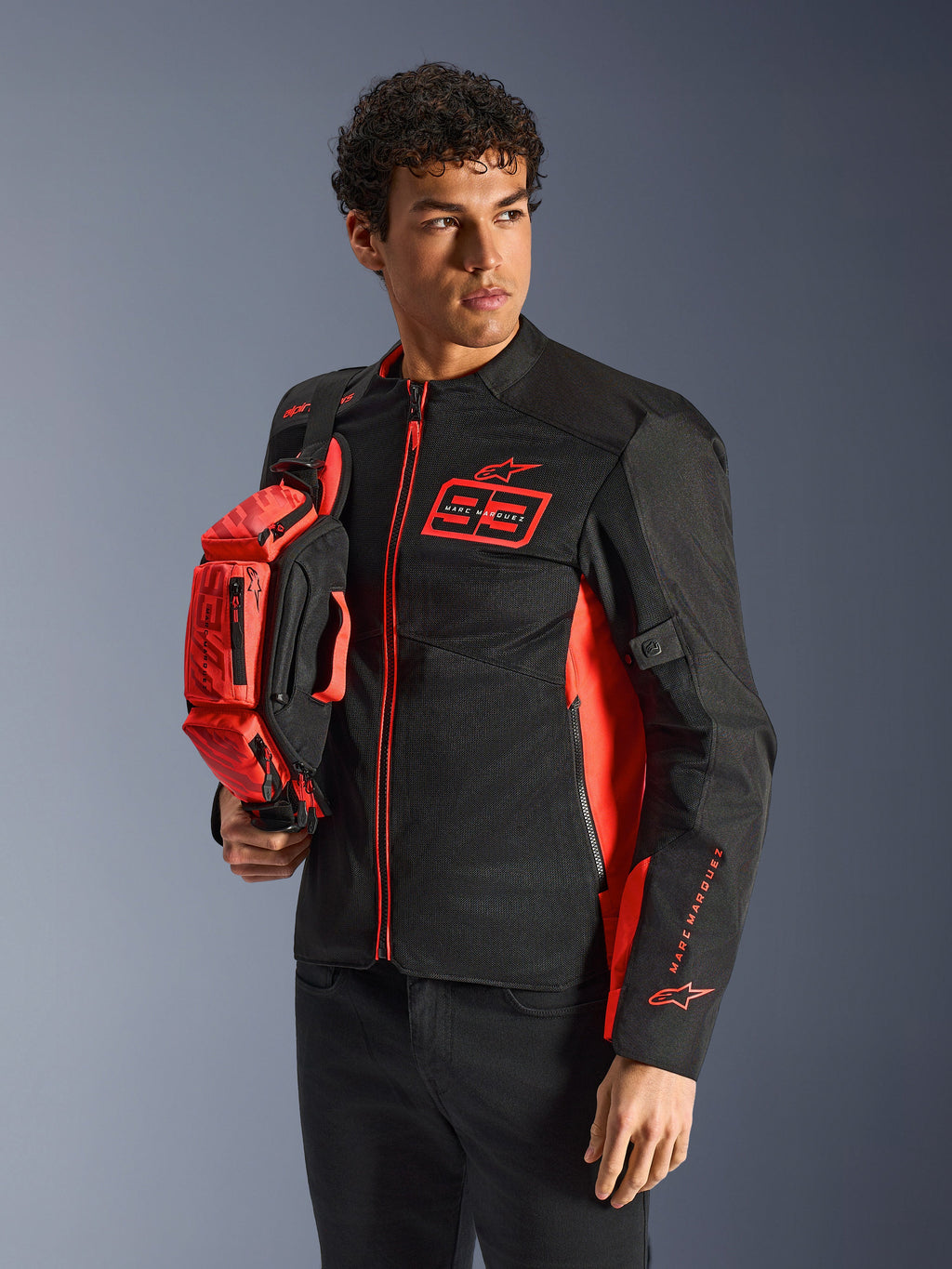 Alpinestars MM93 Waist Bag, Red Fluo and Black, held by a model wearing a matching black and red motorcycle jacket, featuring multiple zippered compartments and Marc Marquez branding, practical gear for sport riding