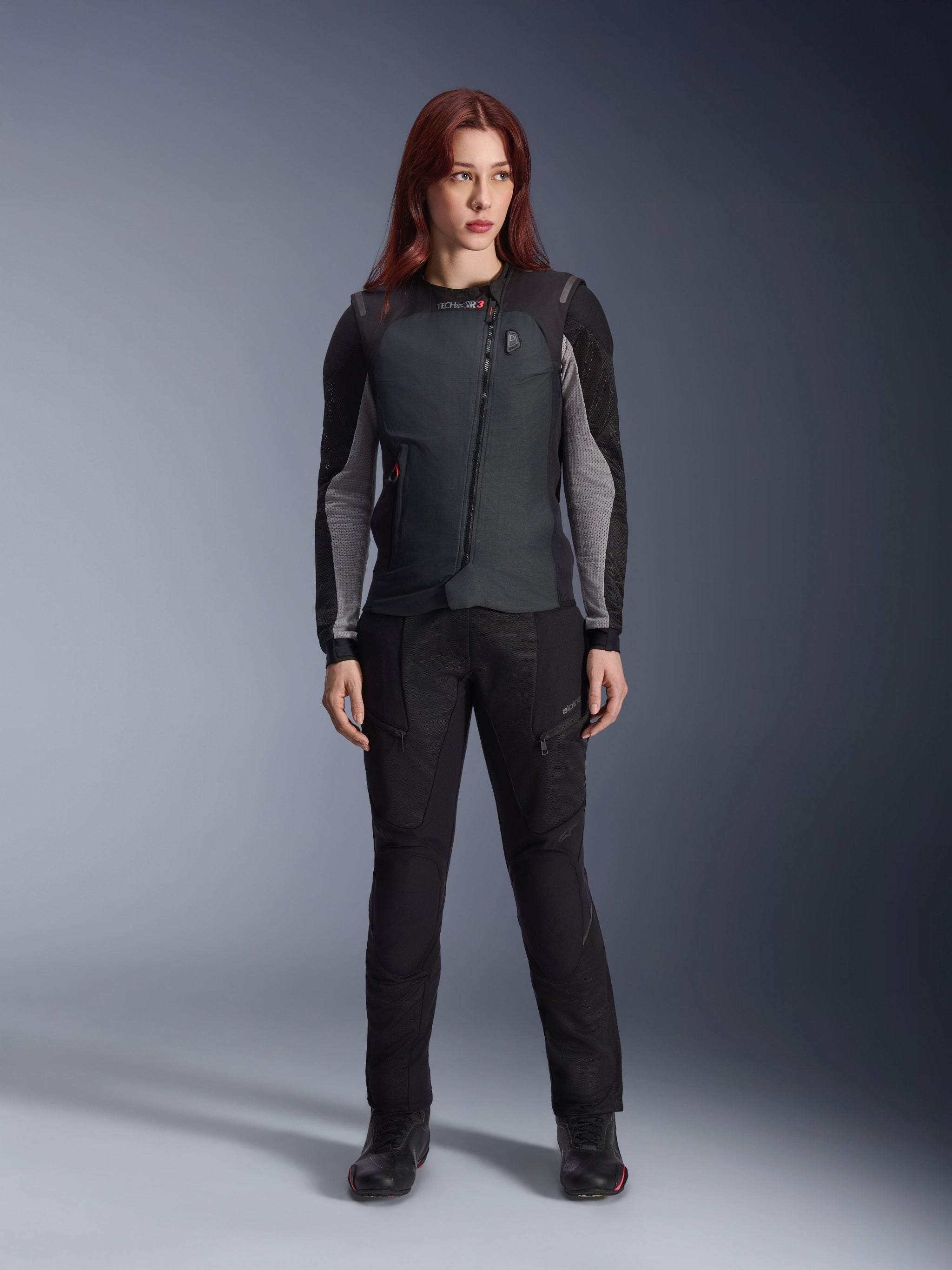 Alpinestars Women Stella Tech-Air® 3 V2 System, Black Motorcycle Airbag Vest, worn over a black and grey long-sleeved technical layer with matching black riding pants, featuring a diagonal zipper and LED status display, tailored female fit for road and touring riders.
