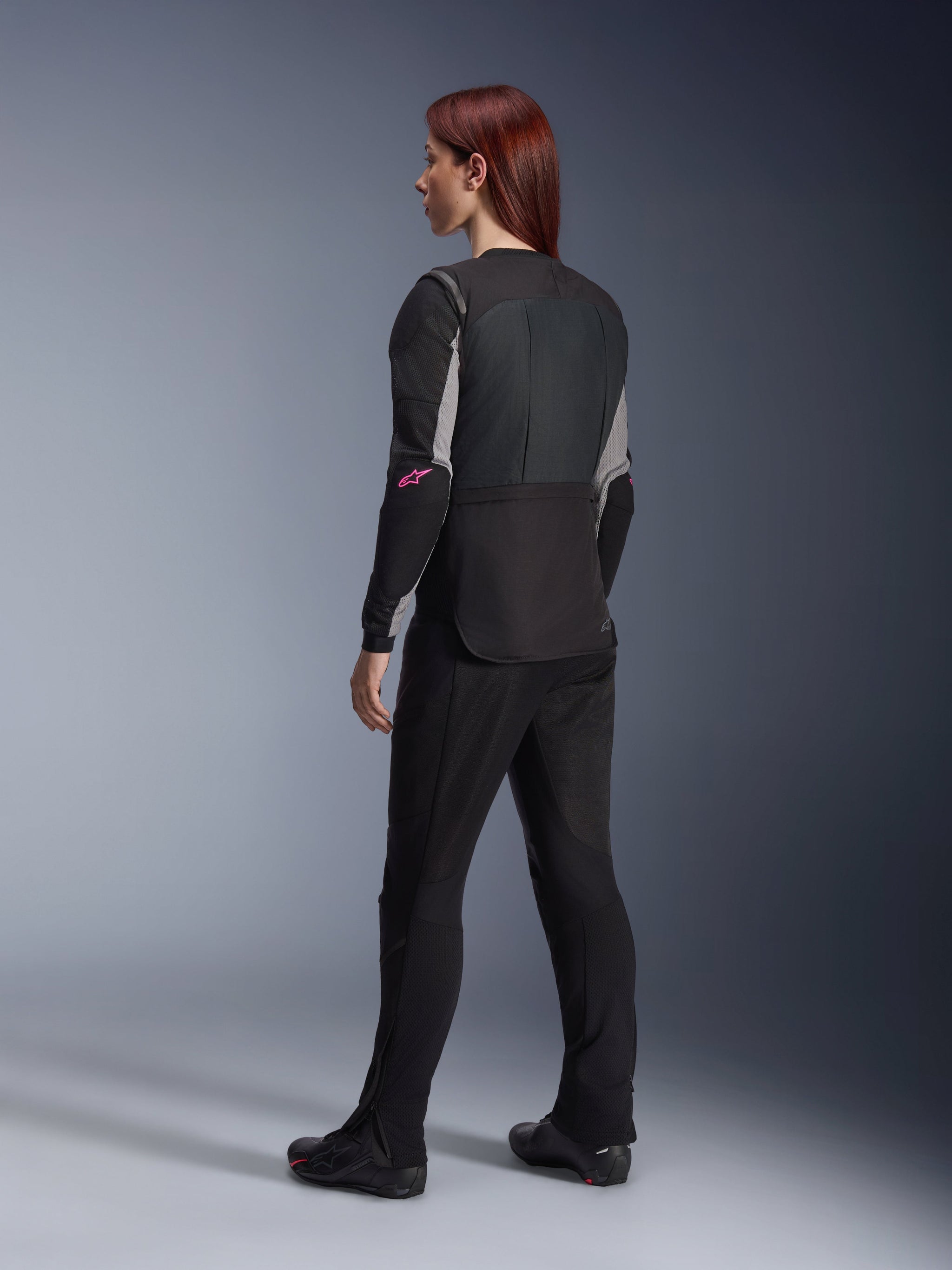 Alpinestars Women Stella Tech-Air® 3 V2 System, Black autonomous airbag vest, back view, worn over a black and grey long-sleeve jersey with pink accents, paired with black motorcycle pants and boots, studio setting