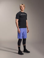 Male model wearing Alpinestars mountain bike apparel, featuring the black A-Aria Switch SS Jersey and Baja Blue A-Aria Lite Shorts, paired with black knee guards, black socks, and black mountain bike shoes in a full-body studio shot.