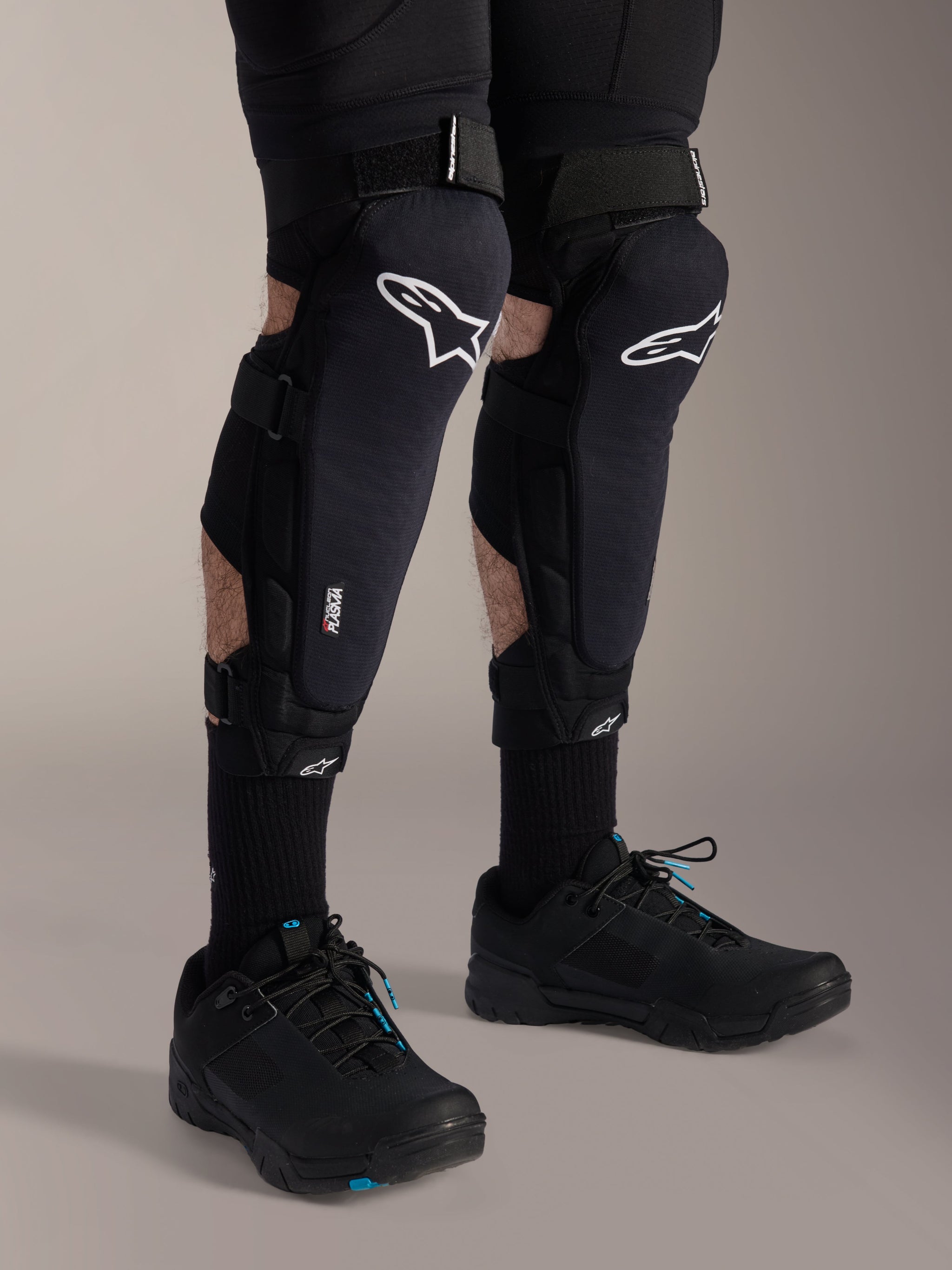 Alpinestars A-Impact Plasma Pro Knee Protectors, Black and White, featuring Nucleon PLASMA technology branding, worn with black socks and black mountain bike shoes, secure hook and loop straps, ergonomic protective gear for off-road cycling