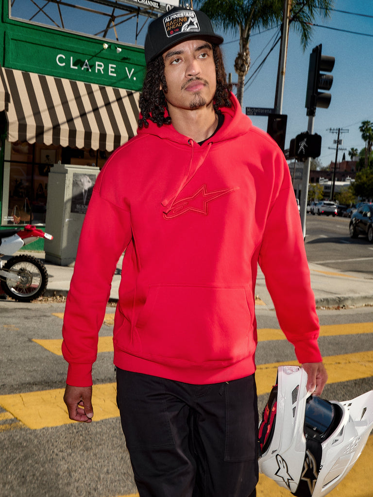 Alpinestars Emergent Oversized Hoodie, Red, 3D embroidered Astars logo on the chest, worn with black pants and a baseball cap while carrying a white motocross helmet, casual urban streetwear style