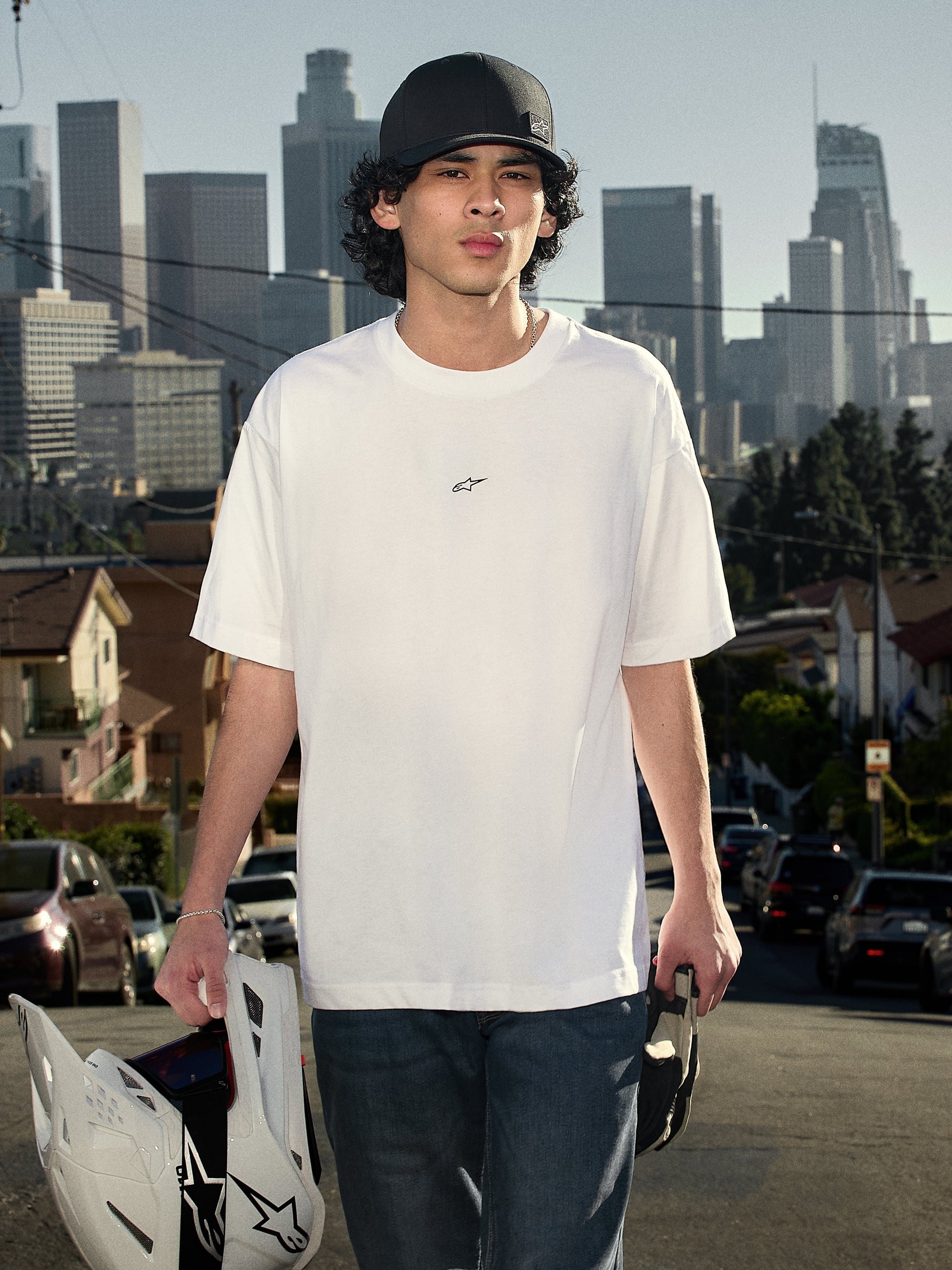 Alpinestars Focused Oversized CSF Tee, Short Sleeve T-Shirt, White, featuring a small black Astars logo on the chest, lifestyle shot of a person wearing the tee and holding a motocross helmet with a city skyline background, casual motorsport streetwear