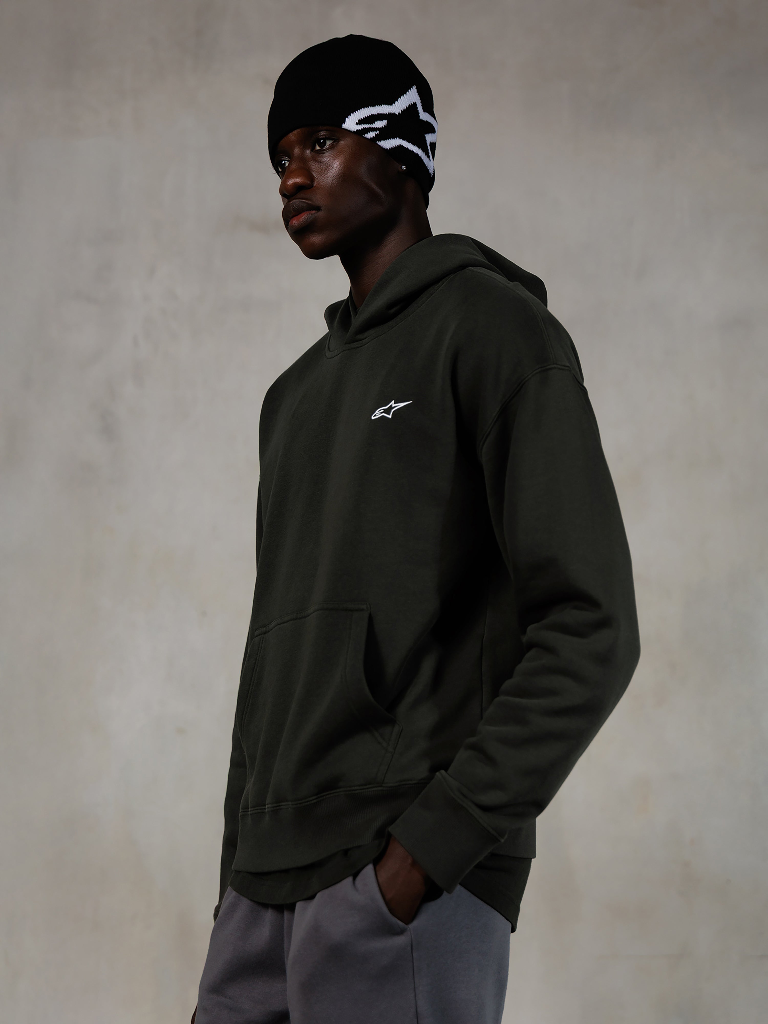 Alpinestars Essentials Ageless Hoodie, Off Black, Male model wearing casual streetwear, dark charcoal stone-washed fleece with a small white star logo, paired with a black Alpinestars beanie and grey pants