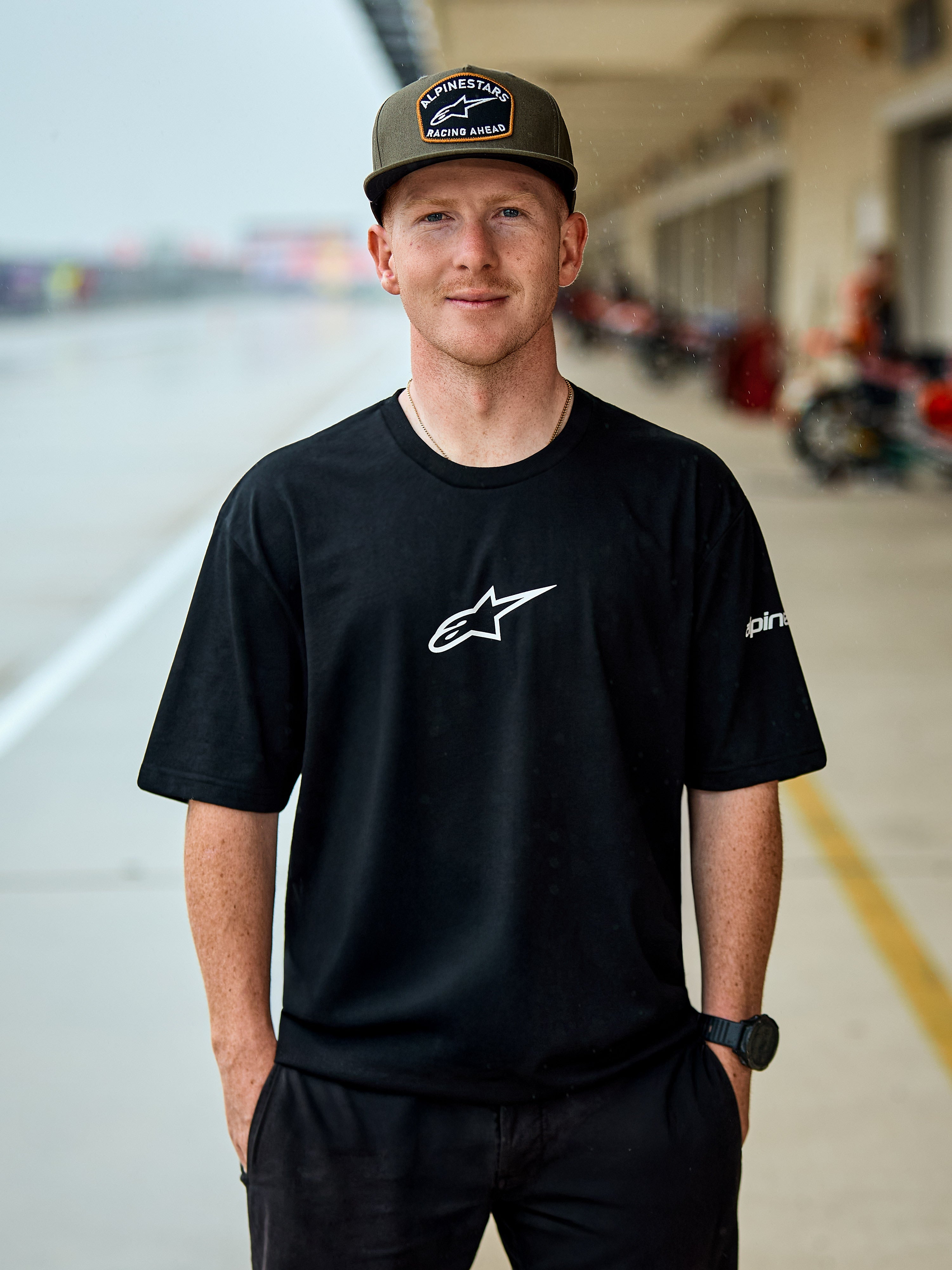 Man wearing Alpinestars Frontal CSF Tee, Short Sleeve T-Shirt, Black, white central Astars logo, olive green Alpinestars cap, casual motorsport-inspired streetwear