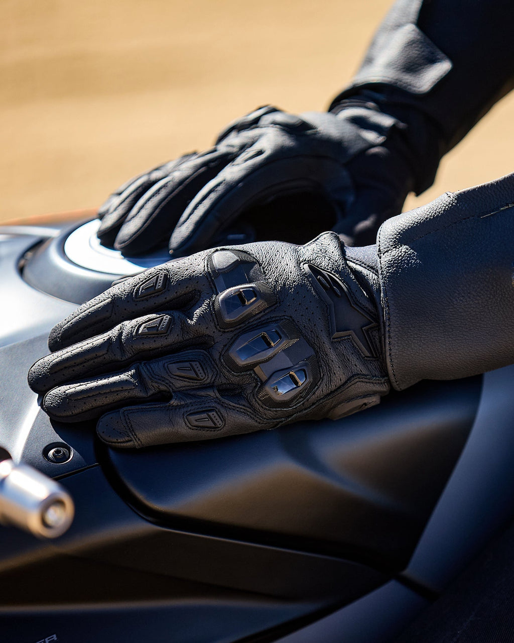 Alpinestars SP X 7 Gloves, Black, short-cuff motorcycle gloves with perforated leather, hands resting on a dark motorcycle fuel tank, featuring visible double-density TPU knuckle protection and embossed branding, designed for sport and road riding.