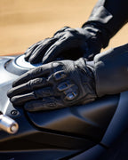 Alpinestars SP X 7 Gloves, Black, short-cuff motorcycle gloves with perforated leather, hands resting on a dark motorcycle fuel tank, featuring visible double-density TPU knuckle protection and embossed branding, designed for sport and road riding.