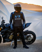 Alpinestars T-Stunt Air Jacket, Sport-Cut Textile Jacket, Black Dark Blue and Bright Blue, worn by a rider standing next to a Ducati motorcycle, featuring a carbon fiber helmet and black riding gear, set against a desert landscape at sunset.