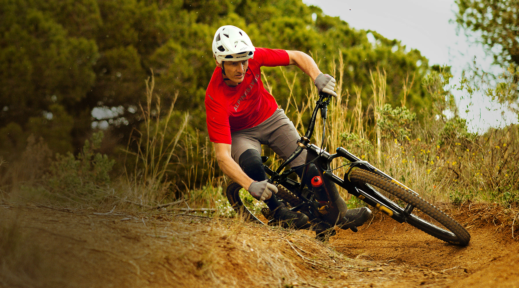 Alpinestars A-Aria Switch Jersey and Vector Tech Solid Helmet, red and white MTB gear on a rider cornering a dirt trail turn.
