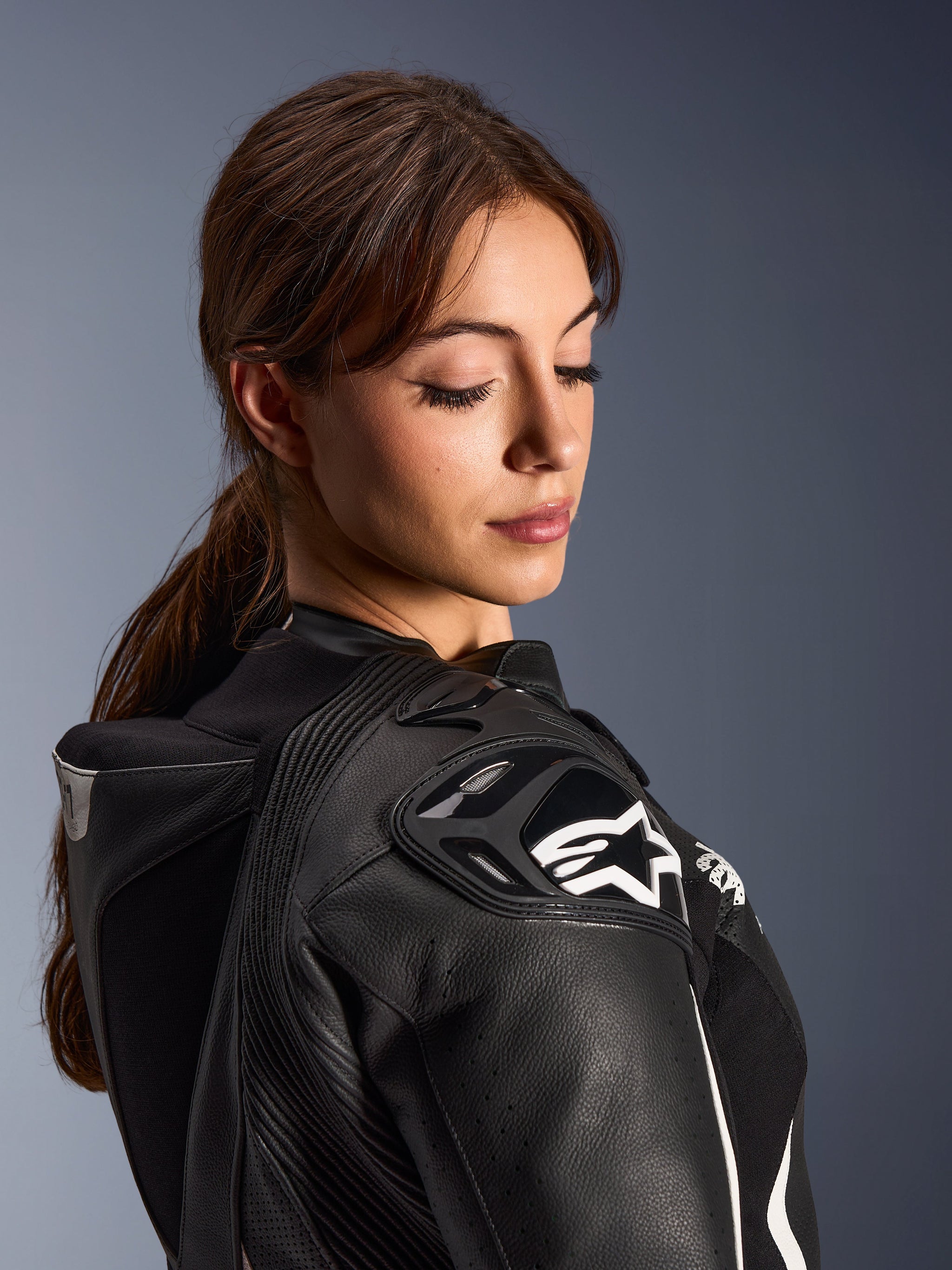 Woman wearing the Alpinestars Stella GP-R7 2-Piece Leather Suit, close-up view of the black leather shoulder with DFS RR armor and white star logo, featuring the integrated aero hump and perforated detailing.