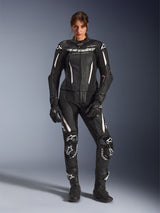 Woman wearing the Alpinestars Stella GP-R7 2-Piece Leather Suit in Black with white accents, featuring perforated leather panels and white branding on the chest and knees, styled with matching black racing gloves and boots, professional motorcycle racing gear